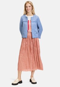 Giacca blu a trama tessuta, top bianco con motivo rosso, gonna midi plissettata rosa e scarpe crema con suole nere e frange.