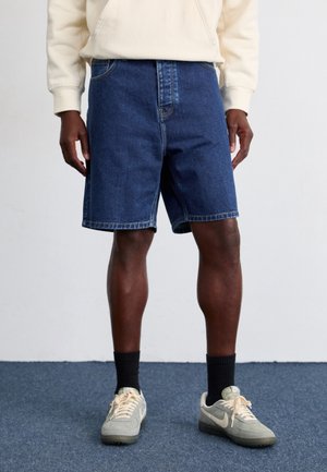 Persona con pantalones cortos de mezclilla azules, calcetines negros, zapatillas beige y una sudadera de color claro, de pie sobre una alfombra azul.