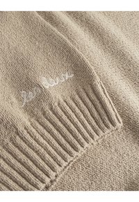 Beige strikket genser med ribbede mansjetter og teksturert finish. Har hvit brodert tekst "les deux" på forsiden.
