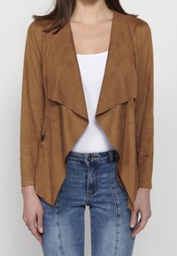 Femme portant une veste marron ouverte façon cascade sur un haut blanc, associée à un jean bleu taille haute, sur un fond uni.