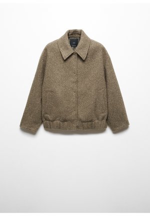 Brun tekstureret uldjakke med krave og trykknapper båret over en hvid sweater og mørke bukser, hånd i lommen.