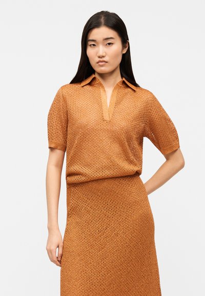 Hosszú, sötét hajú nő, aki texturált, rövid ujjú narancssárga felsőt gallérral és hozzáillő szoknyát visel, egyszerű háttér előtt áll.