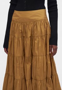 Femme portant une jupe à volants jaune moutarde et un haut noir côtelé à manches longues avec des boutons, les mains reposant sur les côtés.