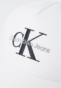 Cappello bianco con visiera curva. Presenta il logo ricamato in nero "CK" e il testo "Calvin Klein Jeans" in grigio. Il tessuto ha una texture liscia.