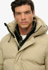 Beige pufferjakke med hætte, med lynlås foran, snoretræk og sorte detaljer. Båret over en grøn skjorte med knapdetaljer.