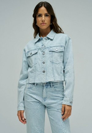 Mujer con chaqueta vaquera de mezclilla azul claro con estampado floral y jeans a juego, posando contra un fondo gris liso.