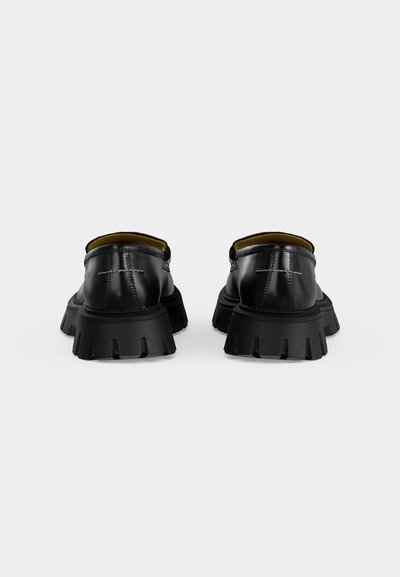 Ζευγάρι μαύρα δερμάτινα loafers με χοντρή σόλα, εμφανιζόμενα από πίσω σε απλό φωτεινό φόντο.