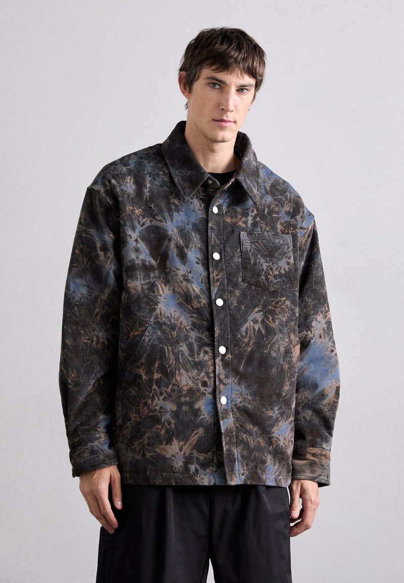 Collared jacket in dark tones featuring a tie-dye pattern of blue and brown. Snap-button closure and a chest pocket with branding.