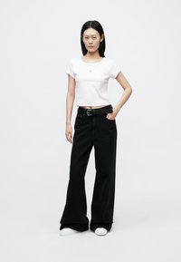 White cropped t-shirt with small red logo, paired with high-waisted black wide-leg jeans and white sneakers. Plain background.