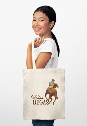 Mladá žena se usmívá a drží tašku na rameno s malbou jezdce na koni a nápisem "Edgar Degas".