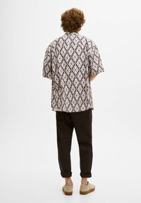 Koszula z krótkim rękawem w kolorze białym z brązowym geometrycznym wzorem; zestawiona z ciemnymi, zwężanymi spodniami i beżowymi butami. Widok z tyłu z detalem na dolnym brzegu.