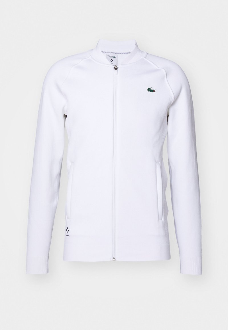 Veste blanche zippée avec manches longues, poignets côtelés et deux poches latérales. Présente un logo vert sur la poitrine et un tissu doux et texturé.