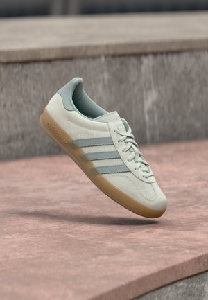 Pantof sport din piele întoarsă bej deschis cu accente albastre, cu trei dungi laterale, talpă texturată de gumă și șireturi plate. Numele modelului "Gazelle" este vizibil.