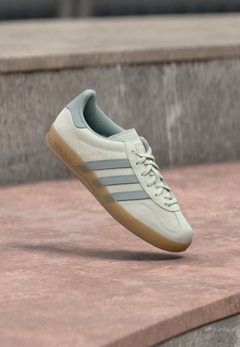 Αθλητικό παπούτσι από μπεζ σουέντ με μπλε λεπτομέρειες, διαθέτει τρεις πλευρικές ρίγες, υφή σόλας από καουτσούκ και επίπεδα κορδόνια. Ονομασία μοντέλου «Gazelle» ορατή.