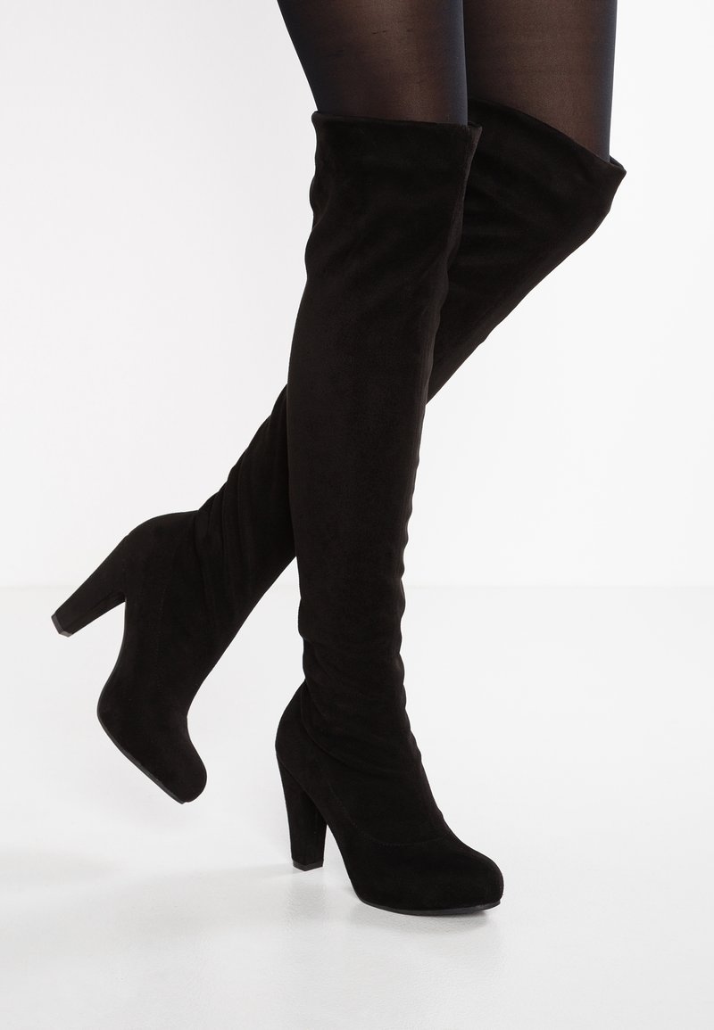 Bottes à talons hauts en daim noir montant au-dessus du genou, portées avec des collants noirs transparents, un pied légèrement relevé sur un fond uni.