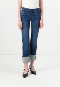 Dark blue denim jeans with a tapered fit, rolled cuffs featuring gray fabric, and gold stitching details. Paired with black pointed shoes.