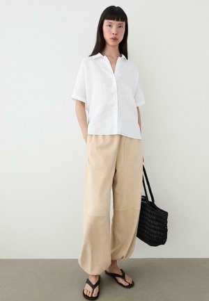 Femme portant une chemise blanche à manches courtes boutonnée, un pantalon beige à jambes larges, des tongs noires, tenant un sac fourre-tout tissé noir sur un fond uni.