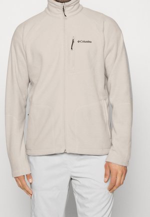 Hellbeige Columbia-Fleecejacke mit durchgehendem Reißverschluss und Brusttasche, getragen von einer Person mit hellgrauer Hose, Hände entspannt an den Seiten.