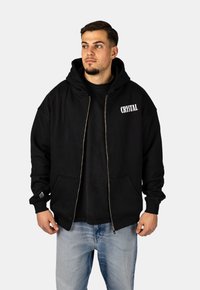 Felpa nera con zip e cappuccio ampio, caratterizzata da un logo frontale "CRYSTAL" in bianco, tessuto strutturato e tasche a marsupio. Indossata sopra una maglietta nera.