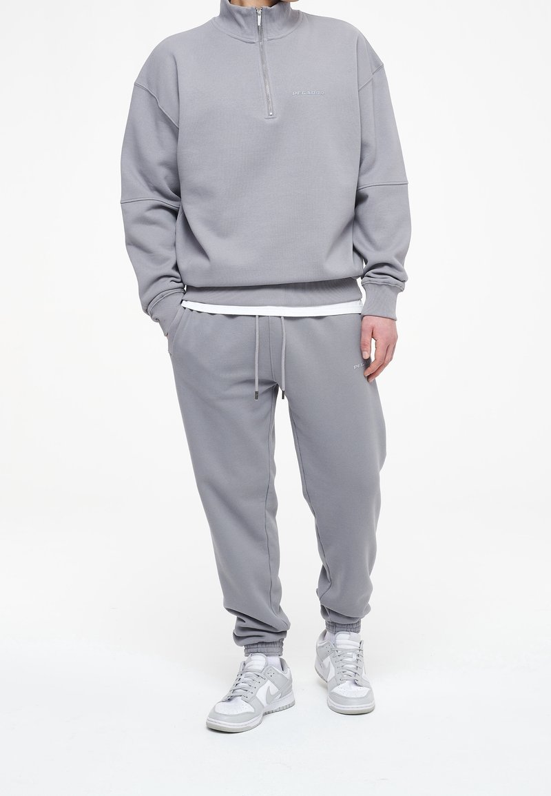 Sudadera gris con cuello de media cremallera y logo, combinada con pantalones jogger a juego. Ambos fabricados en tejido suave con puños de canalé y cinturilla con cordón.