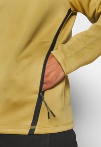 Ręka w kieszeni musztardowej kurtki z czarnym zamkiem błyskawicznym i odblaskowym logo jodełki na rękawie.