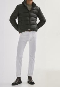 Dunkelgrüne Steppjacke mit horizontalen Nähten und Reißverschluss, kombiniert mit weißen, schlank geschnittenen Hosen und schwarzen Stiefeletten.