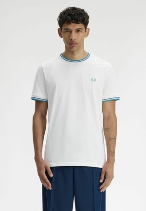 Joven con cabello corto y rizado, que lleva una camiseta blanca con cuello y mangas con rayas azules, y pantalones azul oscuro sobre un fondo liso.