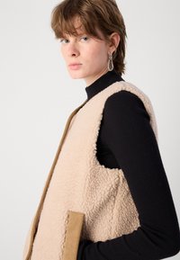 Μπεζ γιλέκο από φλις με απαλή, υφασμάτινη επιφάνεια, διαθέτει πλαϊνή τσέπη και καφέ περιθώριο, φορεμένο πάνω από μαύρο λαιμόκοψη.