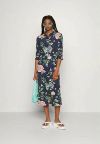 Vestido midi floral em azul-marinho com mangas três quartos, apresentando padrões de flores em rosa e verde. A modelo segura uma bolsa azul-clara e usa sandálias brancas.