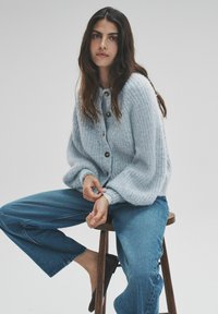 Gaiši zilas krāsas adīts kardigans ar fakturētu apdari, pieciem brūniem pogām, valkāts pār vaļīgām zilām džinsa biksēm uz koka krēsla.