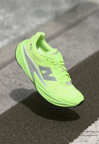 Svijetlo neon zelena New Balance trkaća cipela s reflektirajućim srebrnim logotipom, suspendirana iznad asfaltirane površine s sjenom ispod.