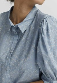 Camicia azzurro chiaro con un motivo testurizzato, colletto classico e maniche a palloncino. Il tessuto appare liscio con una leggera lucentezza e bottoni lungo la parte anteriore.