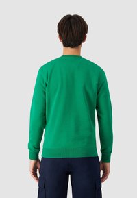 Muž s krátkými tmavými vlasy, oblečený v zeleném svetru s dlouhým rukávem a námořnických cargo kalhotách, stojící zády k jednobarevnému šedému pozadí.