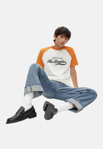 Kurze Raglan-T-Shirt in Weiß mit orangefarbenen Ärmeln, ausgestattet mit bedrucktem Text. Entspannte, hellblaue weit geschnittene Jeanshose und schwarze Slipper.