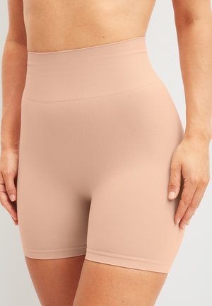 Vrouw die naadloze modelerende shorts met hoge taille in huidkleur draagt, die tot halverwege de dij lopen, tegen een effen achtergrond.