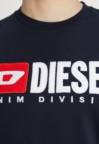Navy blue cotton t-shirt featuring a prominent embroidered logo in red and white, stating "DIESEL" and "DENIM DIVISION," with ribbed collar.