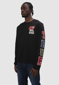 Mann trägt ein schwarzes Langarmshirt mit einem roten Herzen und "MFG. GOOD" auf der Brust sowie großen bunten Buchstaben, die entlang des Ärmels das Wort "TRUE" bilden.