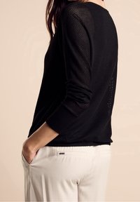 Jersey negro de punto con un patrón texturizado, mangas largas y un cuello redondo, combinado con pantalones de color claro que presentan un detalle de bolsillo trasero.