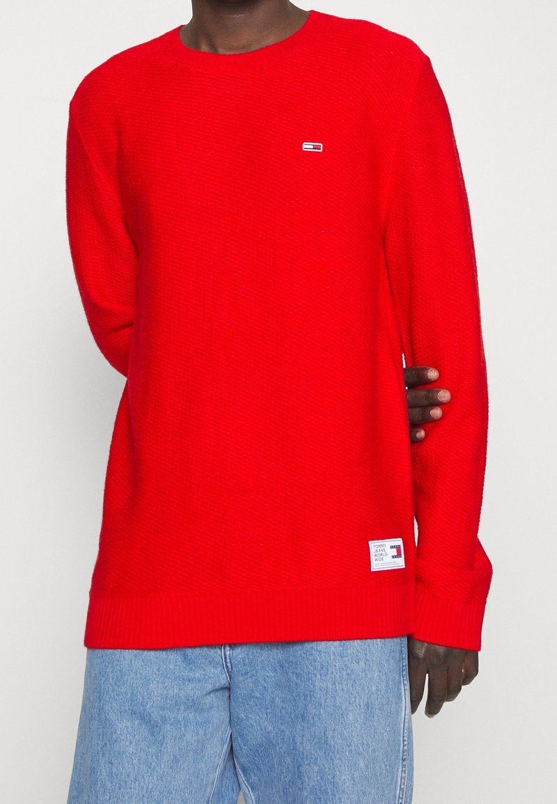 Personne à la peau foncée portant un pull rouge vif à manches longues avec de petits écussons logo et un jean bleu clair, debout devant un arrière-plan uni.