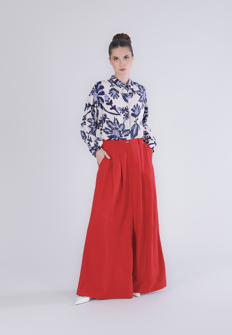 Vrouw staat met handen in de zakken, draagt een witte blouse met blauwe bloemenprint en wijde rode broek, tegen een effen lichte achtergrond.