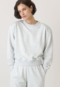 Jasnoszara bluza z miękkiego materiału, z okrągłym dekoltem, ściągaczami przy rękawach i luźnym krojem. Tekstura wydaje się gładka z minimalnymi detalami.