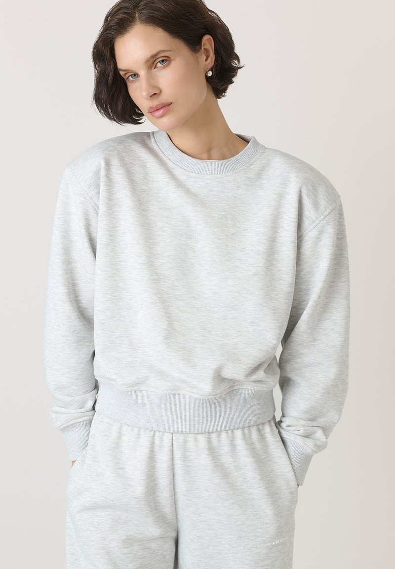 Jasnoszara bluza z miękkiego materiału, z okrągłym dekoltem, ściągaczami przy rękawach i luźnym krojem. Tekstura wydaje się gładka z minimalnymi detalami.