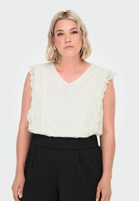 Blond kvinde iført en cremet blonde ærmeløs bluse med bølgede kanter og høj talje sorte bukser, stående mod en ensfarvet baggrund.