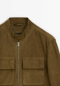 Giacca bomber in suede marrone con colletto alto, zip frontale e due ampie tasche a toppa, caratterizzata da una texture morbida e linee pulite.