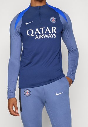 Homme portant un haut de sport bleu et gris à manches longues et un pantalon avec les logos du Paris Saint-Germain et le sponsor Qatar Airways.
