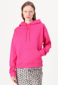 Felpa rosa brillante con tasca frontale e cappuccio con cordino; il tessuto sembra morbido. Indossata sopra una gonna a fantasia floreale.