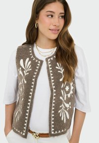 Jeune femme portant une chemise blanche et un gilet texturé marron avec une broderie florale blanche, des boucles d'oreilles anneaux dorés, des colliers dorés superposés et une ceinture marron.