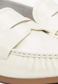 White patent leather loafers featuring a bow design, visible stitching, and a textured around the toe area, with a smooth sole.