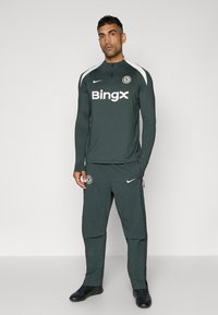 Trening sportiv de culoare verde închis, realizat dintr-un material ușor. Are un guler înalt, mâneci lungi și accente albe cu un logo pe partea de sus și pe pantaloni.