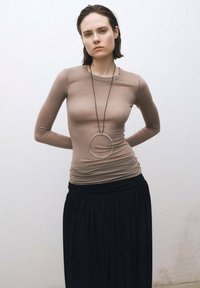 Langärmliges, eng anliegendes beiges Oberteil aus leichtem Stoff, kombiniert mit einem schwarzen Rock. Eine auffällige silberne Rundanhänger hängt an einer feinen Kette.